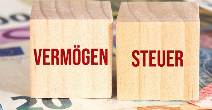 Zwei Holzwürfel, die auf mehreren Euro-Geldscheinen stehen und mit den Wörten Vermögen und Steuer versehen sind