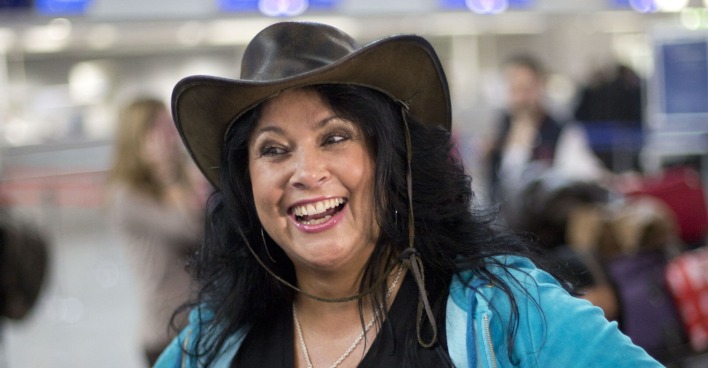 Iris Klein mit einer blauen Weste und einem braunen Cowboyhut auf einem Flughafen.