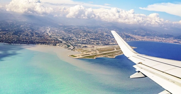 Die Tragfläche eines Flugzeugs aus dem fliegenden Flugzeug fotografiert. Darunter türkises Meer und eine Landebahn