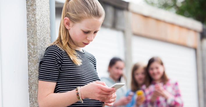 Mädchen schaut traurig in ihr Handy, dahinter stehen lachende Mädchen