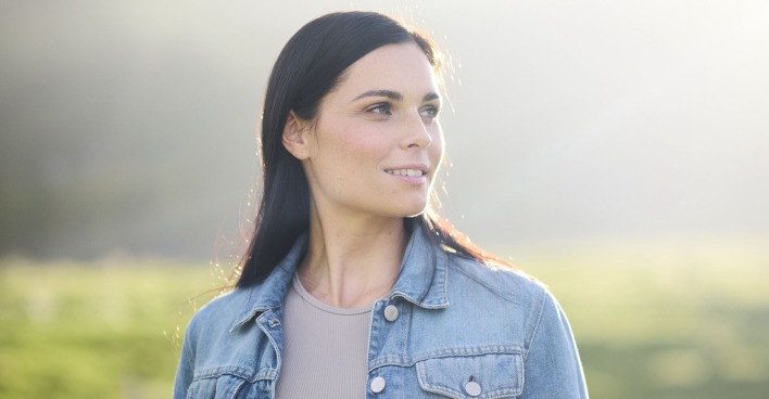 Anna Veith mit offenen schwarzen Haaren und einer blauen Jeansjacke vor einer grünen Wiese.