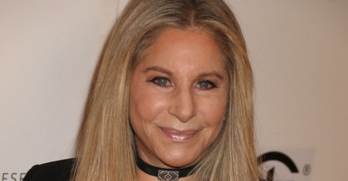 Barbara Streisand mit einer engen schwarzen Kette und offenen blonden Haaren vor einer Fotoleinwand.