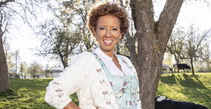Arabella Kiesbauer in einem hellblauen Dirndl mit einer weißen Weste auf einer Kuhwiese.