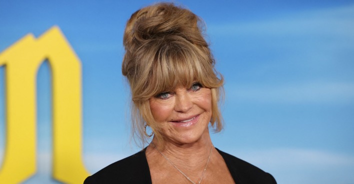 Schauspielerin Goldie Hawn trägt hochgesteckte Haare und steht vor einer blauen Leinwand.