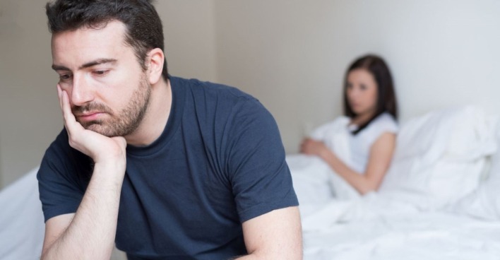 Ein Mann sitzt an den Bettkante, den Kopf in die Hand gestützt. Er wirkt nachdenklich bis bedrückt. Im Bett liegt eine Frau.