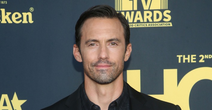 Schauspieler Milo Ventimiglia vor einer schwarzen Fotowand mit einem schwarzen Anzug und gegolten Haaren.