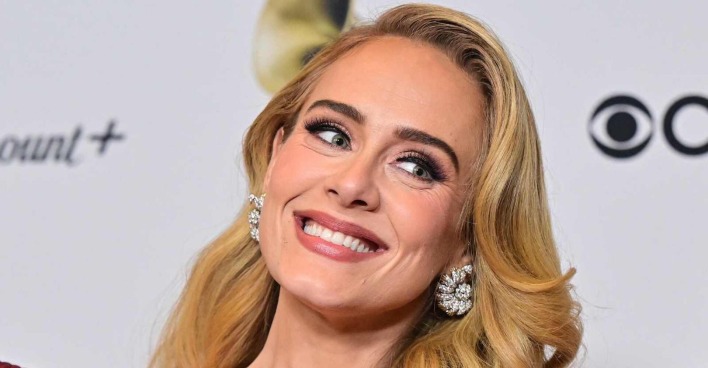Adele trägt eine voluminöse Föhnfrisur und blonde Haare. Sie ist stark geschminkt im Red-Carpet Look und grinst in die Kamera. Sie trägt auffallende Diamantohrringe.