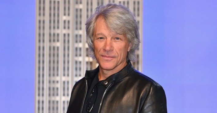 Bon Jovi trägt eine schwarze Lederjacke und steht vor einem hellblauen Hintergrund mit einem Gebäude.