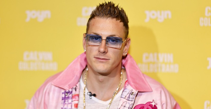 Der Reality Tv Star Calvin Kleinen trägt eine blaue Brille und eine pinke Jacke. Er steht vor einer gelben Fotowand.