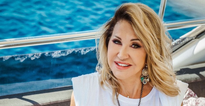 Carmen Geiss hat gewelltes blondes Haar und sitzt auf einer Yacht. Dahinter ist das blaue Meer.