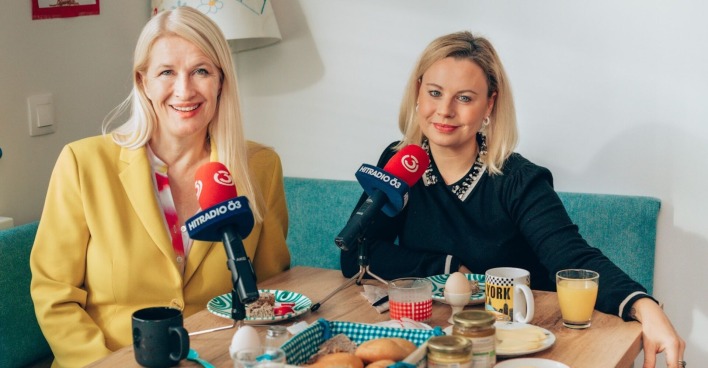 Claudia Stöckl trägt einen gelben Anzug und offene blonde Haare. Katharina Strasser einen dunklen Pullover. Vor ihnen sind zwei rote Mikrofone und ein dicht gedeckter Tisch.