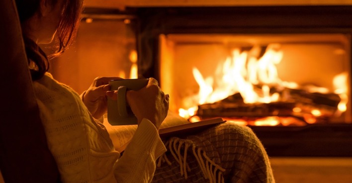Frau sitzt mit Buch vor einem Kamin | Credit: iStock.com/Rasulovs