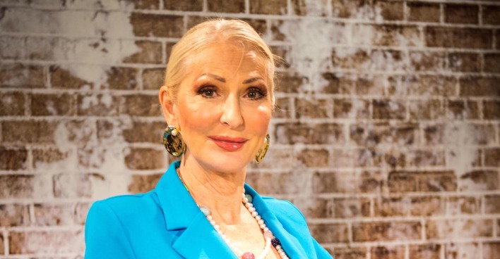 Désirée Nick trägt einen blauen Blazer, zurück gebundene Haare und steht vor einer braunen Ziegelwand.
