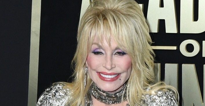 Dolly Parton hat blonde, toupierte Haare, viel Make-up und ein glitzerndes Kleid. Sie steht vor einer schwarzen Fotowand.