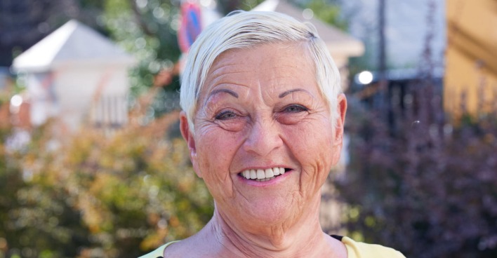 Jazz Gitti steht vor einer Wiese und Häusern. Sie trägt kurze platinblonde Haare und lächelt in die Kamera.