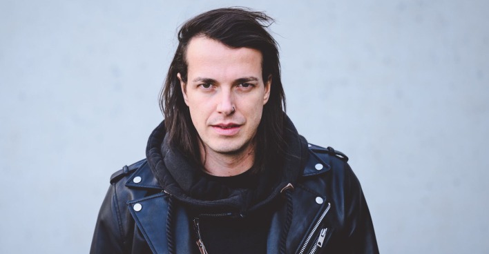 Marco Pogo trägt einen schwarzen Pullover und eine schwarze Lederjacke. Er schaut frontal in die Kamera und steht vor einer grauen Wand.