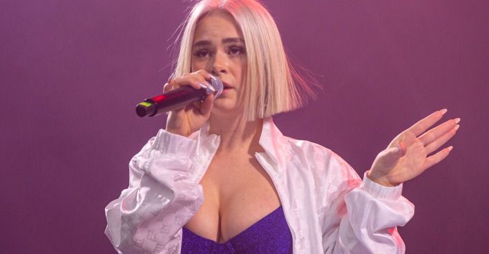 Mathea trägt ein glitzernes Bikinitop und ein weißes Hemd. Sie hat einen blonden Longbob und singt ein Lied.