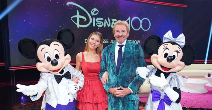 Thomas Gottschalk trägt einen blauen Anzug. Victoria Swarovski ein rotes, kurzes Kleid. Neben ihnen stehen Mickey und Mini Maus.