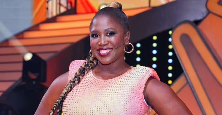 Motsi Mabuse trägt einen schwarzen geflochtenen Zopf und ein silbernes Kleid. Sie lacht in die Kamera und befindet sich vor einem Bühnenbild.