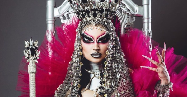 Eine Drag-Queen mit impsanter silvber Krone und pinken Tüll. Sie sitzt auf einem Thron und hält ein Zepter.