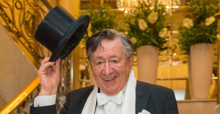 Richard Lugner in der wiener Staatsoper beim Opernball. Er trägt einen schwarzen Anzug und hält einen Hut in die Höhe.
