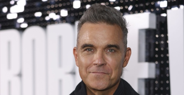 Robbie Williams mit aufgestellten grauen Haaren vor einer Fotoleinwand.