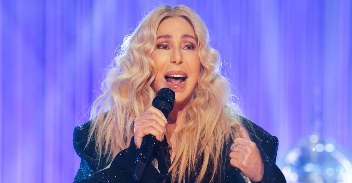 Cher mit blonden, gewellten Haaren und einem schwarzen Sakko. Sie singt in ein Mikrofon.