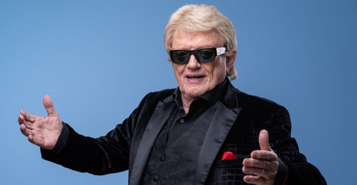 Heino steht vor einer blauen Fotowand mit einem schwarzen Sakko, blonden Haaren und einer Sonnenbrille.