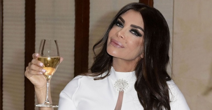 Kader Loth mit einem weißen Kleid und ein Glas Sekt in der Hand. Sie hält den Kopf schief in die Kamera und hat stark geschminkte Augen, sowie lange schwarze Haare.