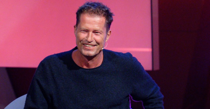 Til Schweiger mit einem dunkelblauen Pullover vor einer Rosinen Filmwand.