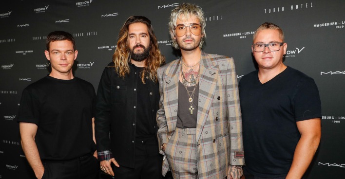 Bill Kaulitz trägt einen karierten Anzug, seine Bandmitglieder sind schwarz angezogen. Sie sehen vor einer schwarzen Fotoleinwand.