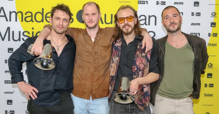 Wanda beim Austrian Musik Award. Sie tragen lässige Kleidung, halten zwei Megafone und stehen vor einer Fotowand auf der steht "Amadeus Music Award".