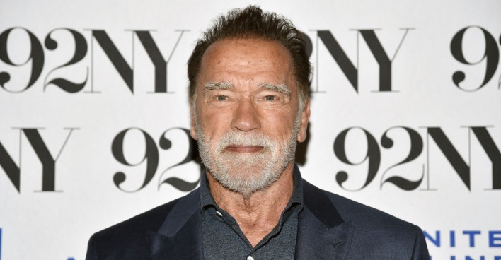 Arnold Schwarzenegger mit einem dunkelgrauen Smoking vor einer Fotowand.