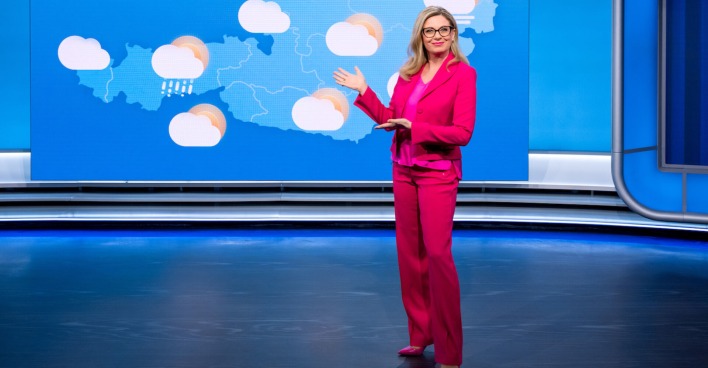 Christa Kummer in einem pinken Hosenanzug vor einer Wetterwand.
