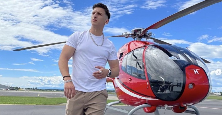 Satans Bratan mit einem weißen T-Shirt vor einem roten Helikopter.