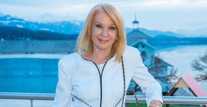 Gerda Rogers auf einem Balkon. Hinter ihr ist ein See und Berge. Sie trägt eine weiße Jacke und offene blonde Haare.