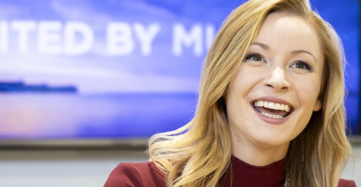 Kaleen trägt offene blonde Haare und einen roten Rollkragenpullover.