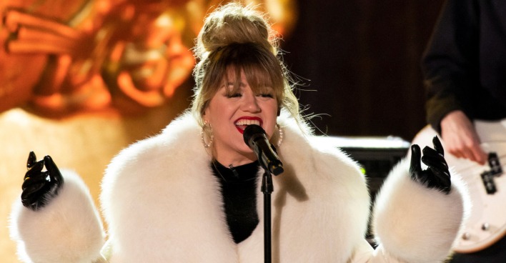 Kelly Clarkson trägt einen weißen Pelzmantel und steht auf einer Bühne.