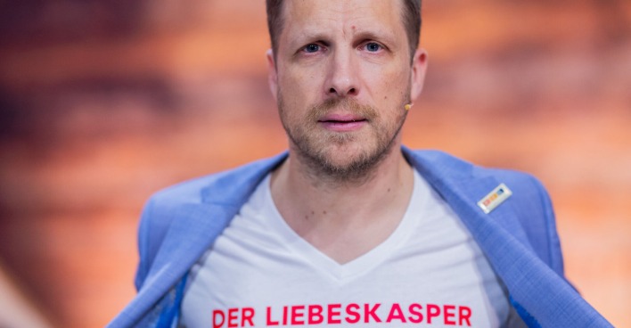 Oliver Pocher mit einem weißen T-Shirt auf dem steht "Liebeskasper" und einem hellblauen Sakko.