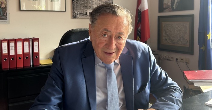 Richard Lugner auf seinem Schreibtisch. Er trägt einen blauen Anzug und eine Krawatte.