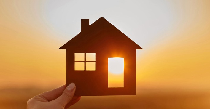 Junge Frau hält ein Miniaturhaus gegen das Licht der untergehenden Sonne | Credit: iStock.com/bodnarchuk