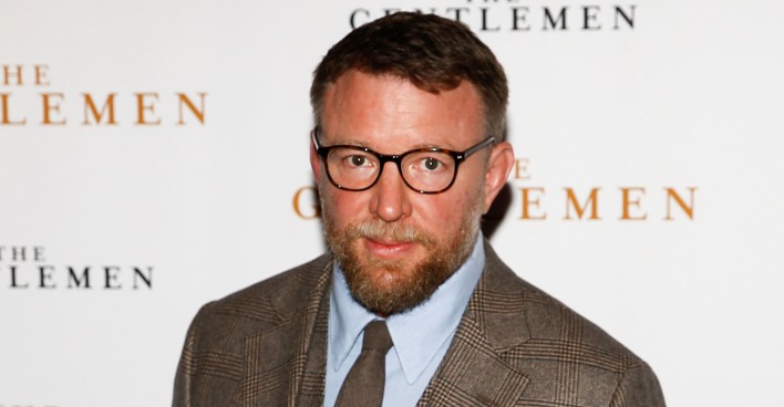 Guy Ritchie vor einer Fotoleinwand mit einem beigen Sakko.
