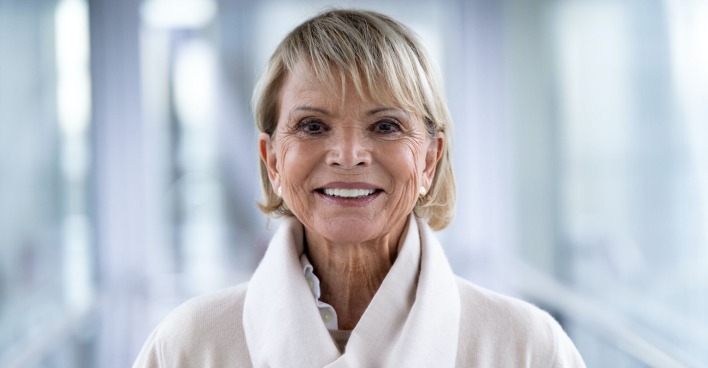 Uschi Glas mit einem weißen Schal und einem beigen Pullover. Sie lacht in die Kamera.