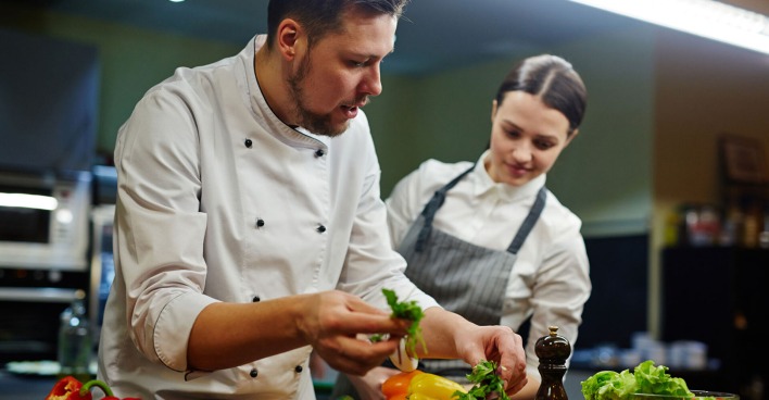 Koch bringt einem weiblichen Lehrling bei, wie man ein Gericht appetitlich anrichtet | Credit: iStock.com/shironosov