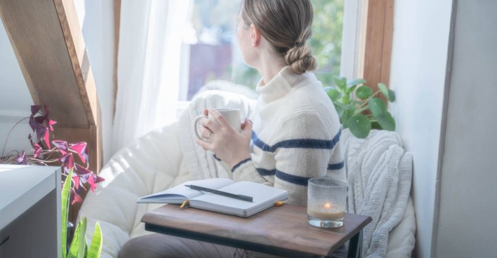 Junge Frau mit Blick aus dem Fenster und aufgeschlagenem Notizbuch auf dem Schreibtisch.