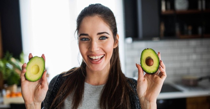 Eine hübsche Frau posiert mit je einer aufgeschnittenen Avocadohälfte, die sie lächelnd in die Kamera hält