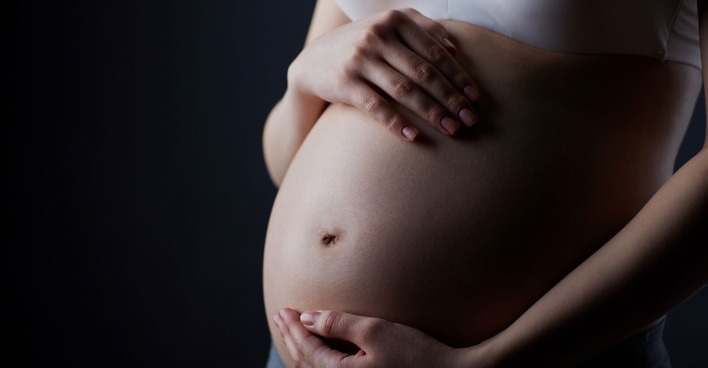 Eine Frau hält zärtlich ihren schwangeren Bauch mit beiden Händen, die Grundstimmung ist düster.