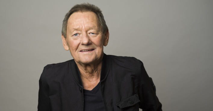 Wolfgang Ambros mit einem schwarzen Outfit vor einer grauen Fotowand.