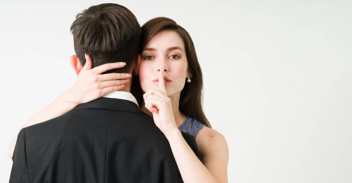 Junge Frau, die ihre Hand um den Nacken ihrer Affäre gelegt hat, der mit dem Rücken zur Kamera steht. Sie hat den ausgestreckten Zeigefinger auf die Lippen gelegt, um ein "Geheimnis" zu symbolisieren