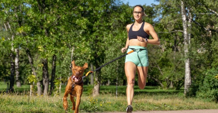 Junge Frau mit schwarzem Top, türkisfarbenen Shorts und Hund mit Canicross-Geschirr um die Hüfte